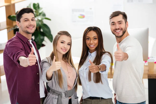 Retrato de jovens sorridentes multirraciais pessoas de negócios mostrando polegares para cima e olhando para a câmera no escritório — Fotografia de Stock