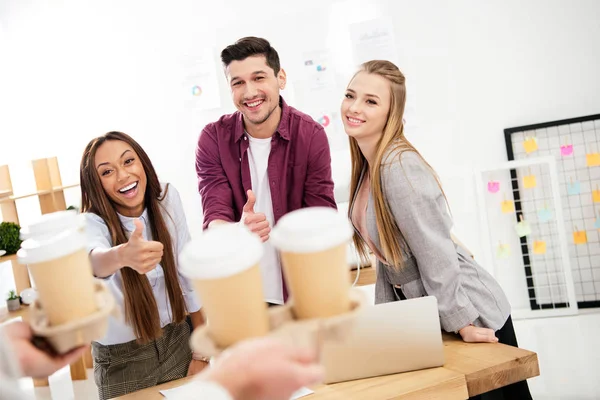 Visión parcial de empresario trajo café para ir para colegas multiétnicos con pulgares en la oficina - foto de stock