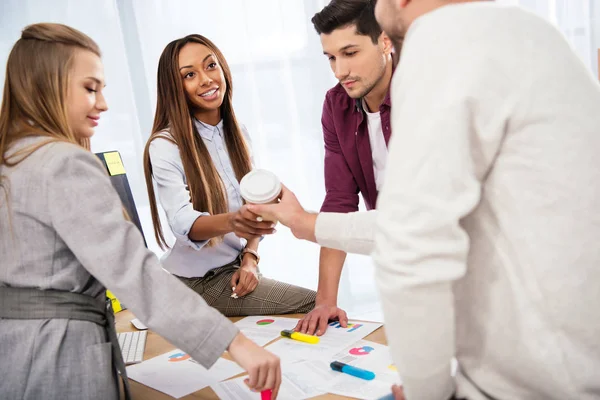 Selective focus of businessman giving coffee to go to african american businesswoman during meeting with colleagues in office — Stock Photo