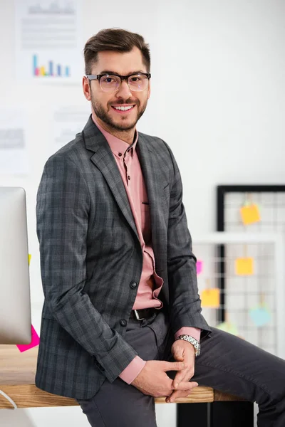 Retrato de empresário sorridente de terno e óculos sentado na mesa e olhando para a câmera no escritório — Fotografia de Stock