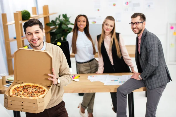 Enfoque selectivo de sonriente hombre de negocios con pizza en las manos y colegas multiétnicos detrás en la oficina - foto de stock