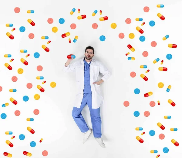 Collage creativo de guapo joven médico acostado en blanco rodeado de varias píldoras - foto de stock