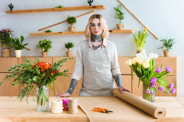 Beautiful young florist in apron and eyeglasses standing at workplace and looking at camera — Stock Photo