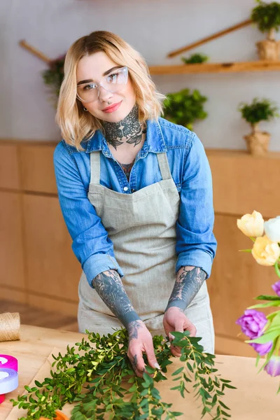 Beautiful young florist in eyeglasses arranging plants and smiling at camera in flower shop — Stock Photo