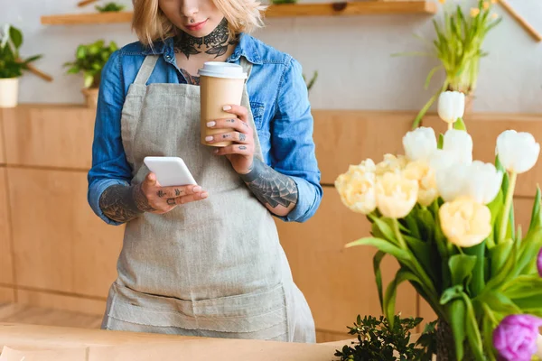 Plan recadré de jeune fleuriste tenant tasse en papier et en utilisant smartphone dans le magasin de fleurs — Photo de stock