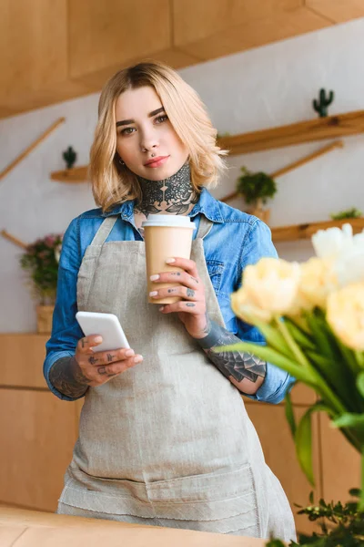 Schöne junge Floristin hält Coffee to go und Smartphone in der Hand und blickt in die Kamera im Blumenladen — Stockfoto