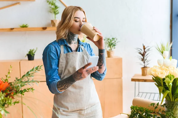 Молодий флорист в фартусі п'є каву з паперової чашки і використовує смартфон у квітковому магазині — стокове фото
