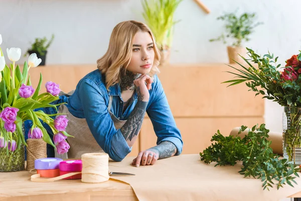 Florista joven pensativo apoyado en la mesa y mirando hacia otro lado en la florería - foto de stock