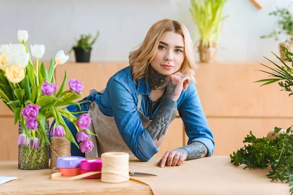 Schöne junge Floristin lehnt am Tisch und blickt in die Kamera im Blumenladen — Stockfoto