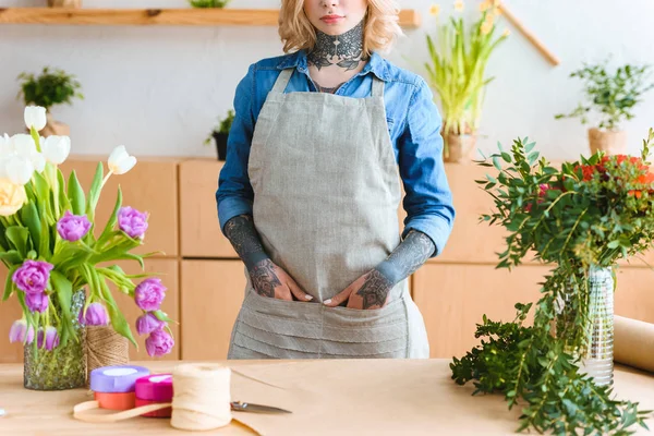 Cropped shot of young female florist in apron standing with hands in pockets in flower shop — Stock Photo