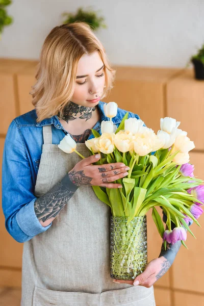 Beautiful young woman in apron holding vase with tulips in flower shop — Stock Photo