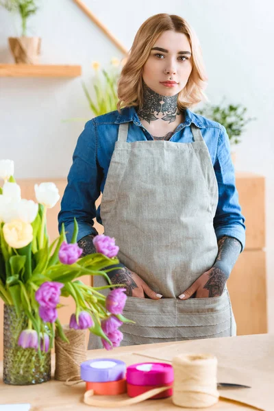 Schöne junge Floristin in Schürze blickt in Blumenladen in die Kamera — Stockfoto