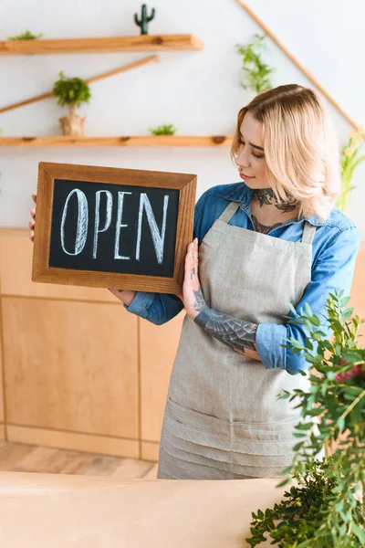 Belle jeune femme dans tablier tenant signe ouvert dans la boutique de fleurs — Photo de stock