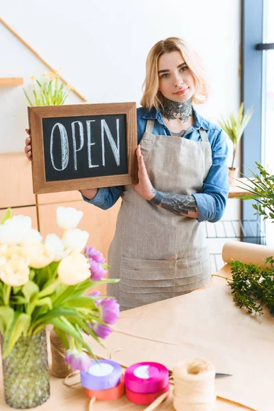 Beautiful young florist holding open sign and looking at camera in flower shop — Stock Photo