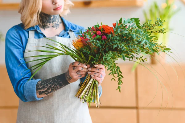 Tiro recortado de mujer joven tatuada en delantal sosteniendo hermoso ramo en la tienda de flores - foto de stock