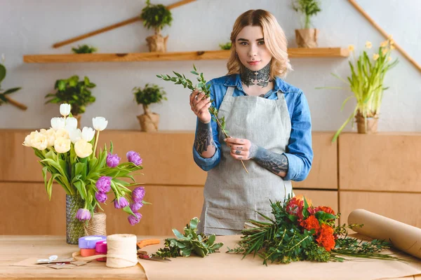 Beautiful young florist in apron holding green plants and looking at camera at workplace — Stock Photo