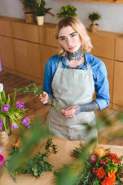 Schöne junge Floristin in Schürze und Brille, die Blumen arrangiert und in die Kamera schaut — Stockfoto