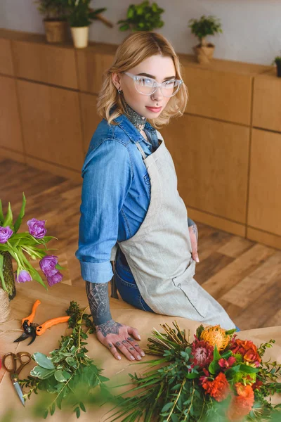 Belle jeune femme en lunettes debout sur le lieu de travail et regardant la caméra — Photo de stock