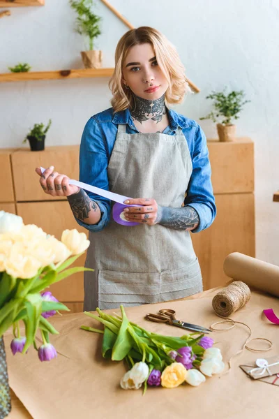 Floristería sosteniendo la cinta y mirando a la cámara mientras trabaja en la floristería - foto de stock