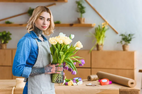 Florista segurando vaso com flores de tulipa e olhando para a câmera — Fotografia de Stock