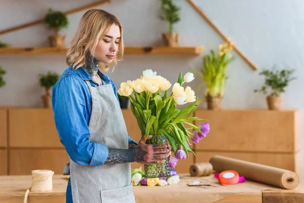 Schöne junge Floristin in Schürze hält Vase mit Tulpenblumen — Stockfoto