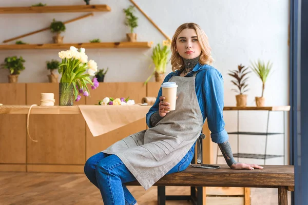 Pensive young florist holding paper cup and looking away in flower shop — Stock Photo