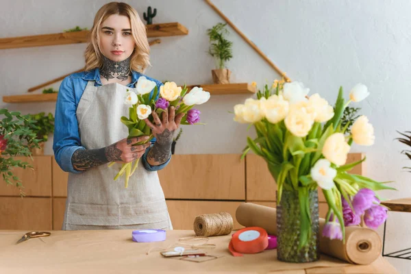 Florista joven en delantal sosteniendo hermosos tulipanes y mirando a la cámara en la tienda de flores - foto de stock