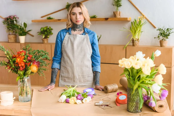 Hermosa joven florista femenina con tatuajes mirando a la cámara en la tienda de flores - foto de stock