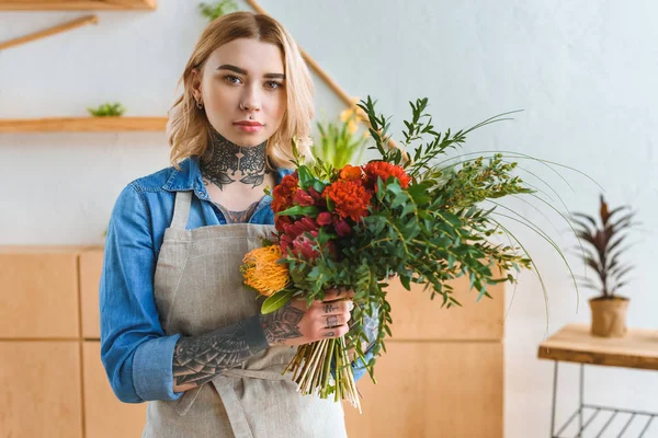 Hermosa florista joven con tatuajes con ramo de flores y mirando a la cámara - foto de stock