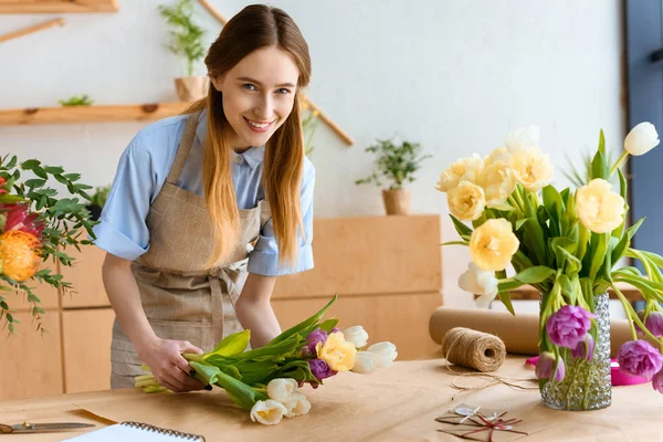 Hermosa florista joven envolviendo tulipanes en papel artesanal y sonriendo a la cámara - foto de stock