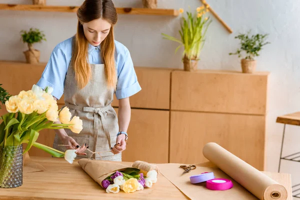 Beautiful young florist in apron arranging flower bouquet at workplace — Stock Photo