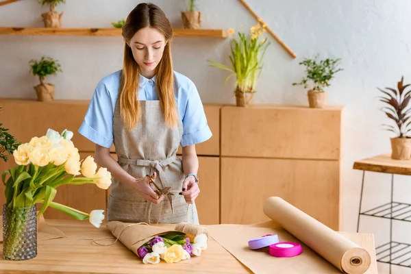 Young florist in apron arranging flower bouquet at workplace — Stock Photo