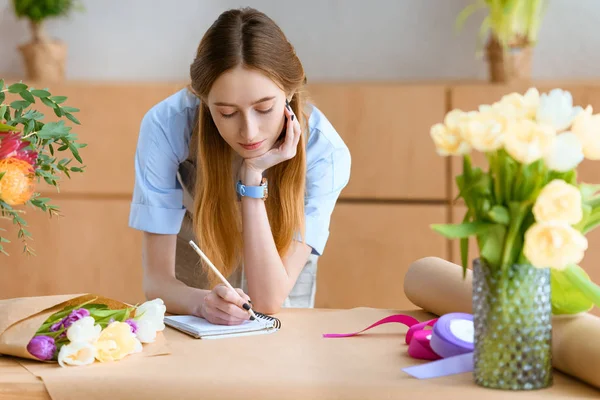 Schöne junge Floristin macht sich Notizen in Notizbuch am Arbeitsplatz — Stockfoto