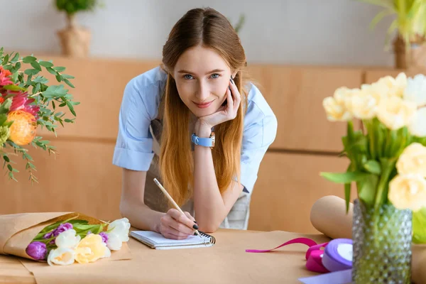 Beautiful young florist taking notes and smiling at camera at workplace — Stock Photo