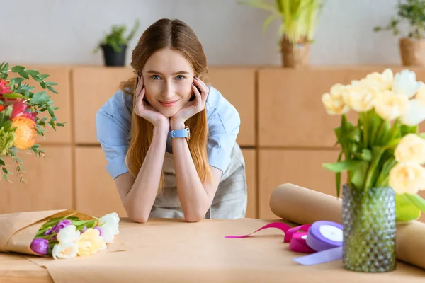 Beautiful young florist leaning at table with flowers and craft paper and smiling at camera — Stock Photo