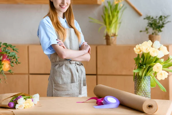 Cropped shot of smiling young florist standing with crossed arms in flower shop — Stock Photo