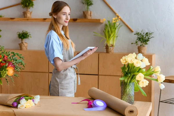 Schöne lächelnde junge Floristin, die ein digitales Tablet in der Hand hält und im Blumenladen wegschaut — Stockfoto