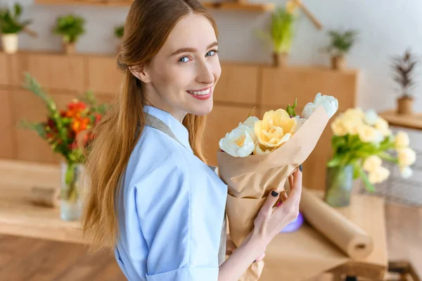 Beautiful young female florist holding bouquet of tulips and smiling at camera in flower shop — Stock Photo