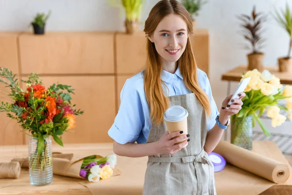 Bela jovem florista segurando smartphone e café para ir, sorrindo para a câmera na loja de flores — Fotografia de Stock