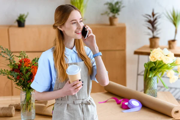Sourire jeune fleuriste tenant du café pour aller et parler par smartphone — Photo de stock