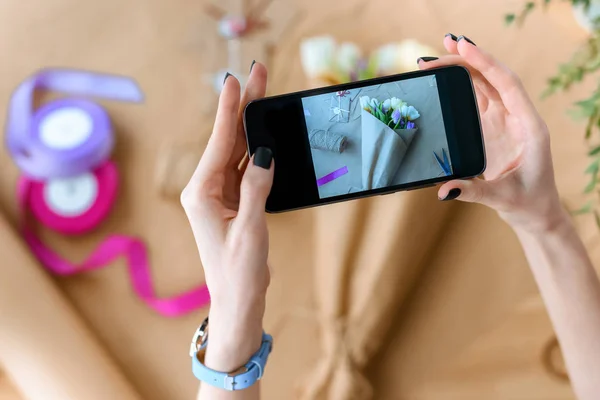 Cropped shot of woman holding smartphone and photographing beautiful bouquet of tulips in craft paper — Stock Photo