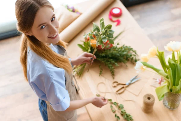 Vue grand angle de beau jeune fleuriste arrangeant des plantes et souriant à la caméra — Photo de stock