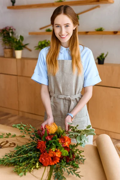 Hermosa florista joven arreglando ramo de flores y sonriendo a la cámara - foto de stock