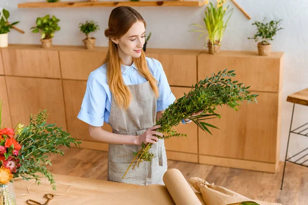 Vue grand angle du jeune fleuriste souriant dans le tablier tenant des plantes vertes dans le magasin de fleurs — Photo de stock