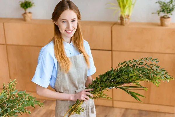 Bela jovem florista feminina segurando plantas verdes e sorrindo para a câmera na loja de flores — Fotografia de Stock