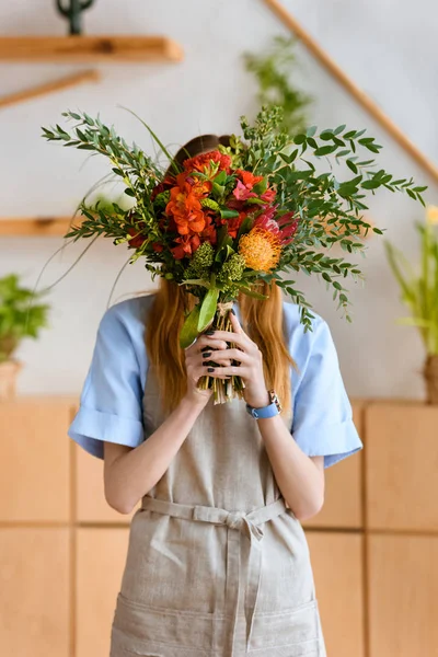 Junge Floristin in Schürze versteckt Gesicht hinter schönem Blumenstrauß — Stockfoto