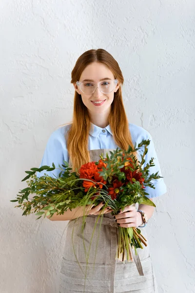 Hermosa florista joven en gafas hoding ramo de flores y sonriendo a la cámara - foto de stock