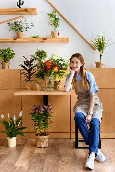 Schöne junge Floristin lächelt in die Kamera, während sie im Blumenladen sitzt — Stockfoto