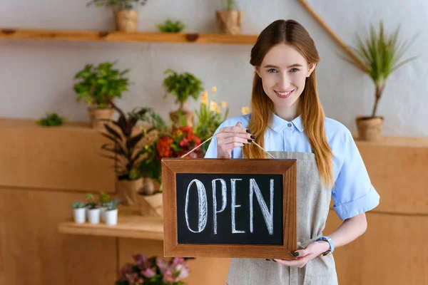 Beautiful young florist in apron holding sign open and smiling at camera in flower shop — Stock Photo