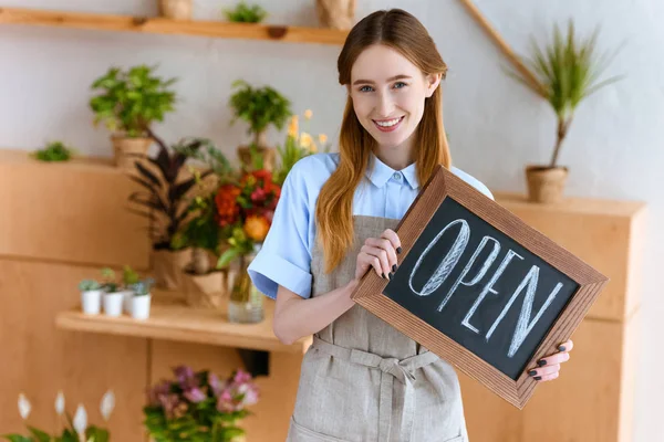 Hermosa joven en delantal sosteniendo el cartel abierto y sonriendo a la cámara en la tienda de flores - foto de stock